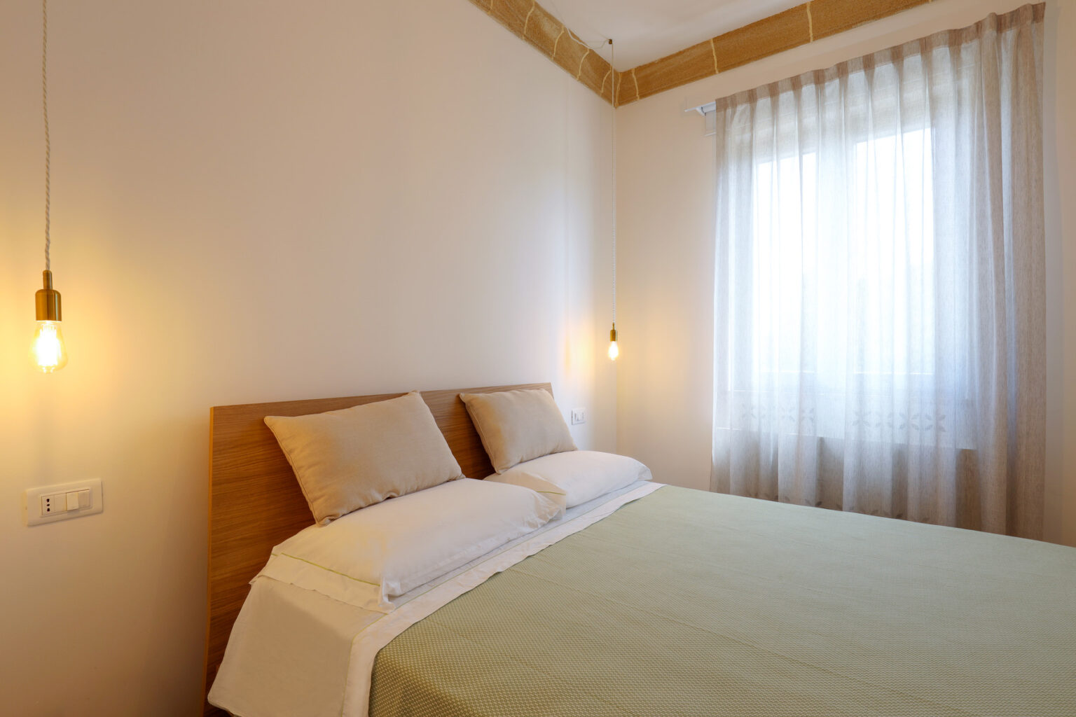 Cozy bedroom interior featuring a wooden bed frame with soft pillows, a light green bedspread, and warm pendant lighting. Natural light filters through sheer curtains, creating a serene atmosphere. Ideal for home decor inspiration. | Only on SopranoVillas