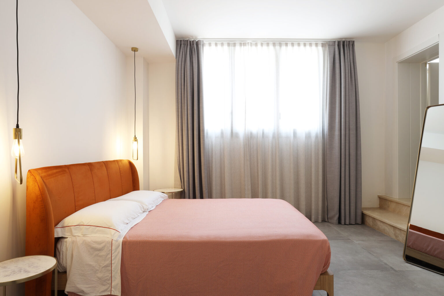 Modern bedroom interior featuring a cozy orange headboard, a pink bedspread, large windows with sheer curtains, and a stylish full-length mirror. The minimalist design emphasizes natural light and a calming atmosphere. | Only on SopranoVillas