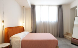 Modern bedroom interior featuring a cozy orange headboard, a pink bedspread, large windows with sheer curtains, and a stylish full-length mirror. The minimalist design emphasizes natural light and a calming atmosphere.