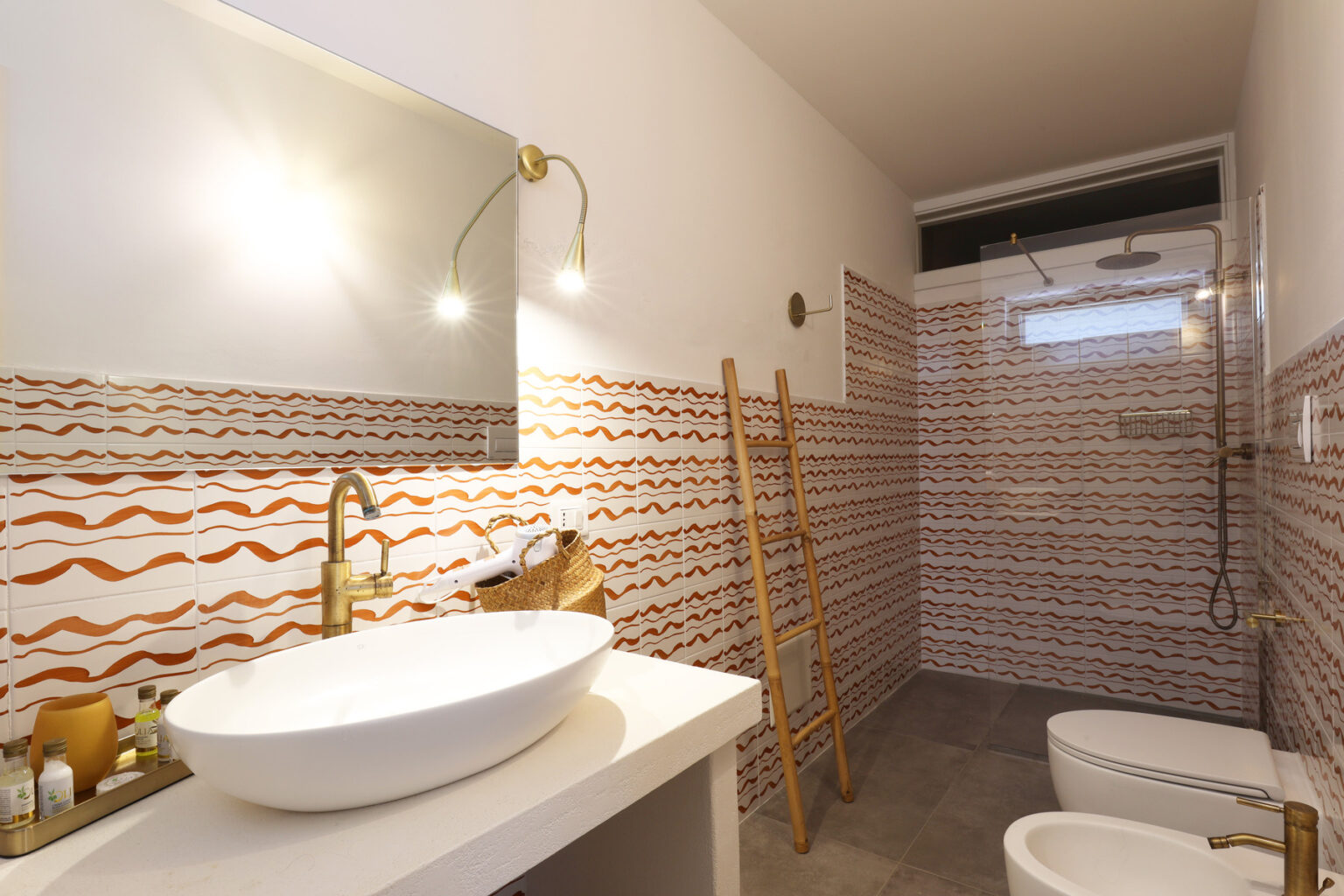 Modern bathroom design featuring a stylish white sink, gold faucet, and decorative wave-patterned tiles in warm tones. The space includes a shower area and a wooden ladder for storage, creating a contemporary and functional aesthetic. | Only on SopranoVillas