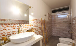 Modern bathroom design featuring a stylish white sink, gold faucet, and decorative wave-patterned tiles in warm tones. The space includes a shower area and a wooden ladder for storage, creating a contemporary and functional aesthetic.