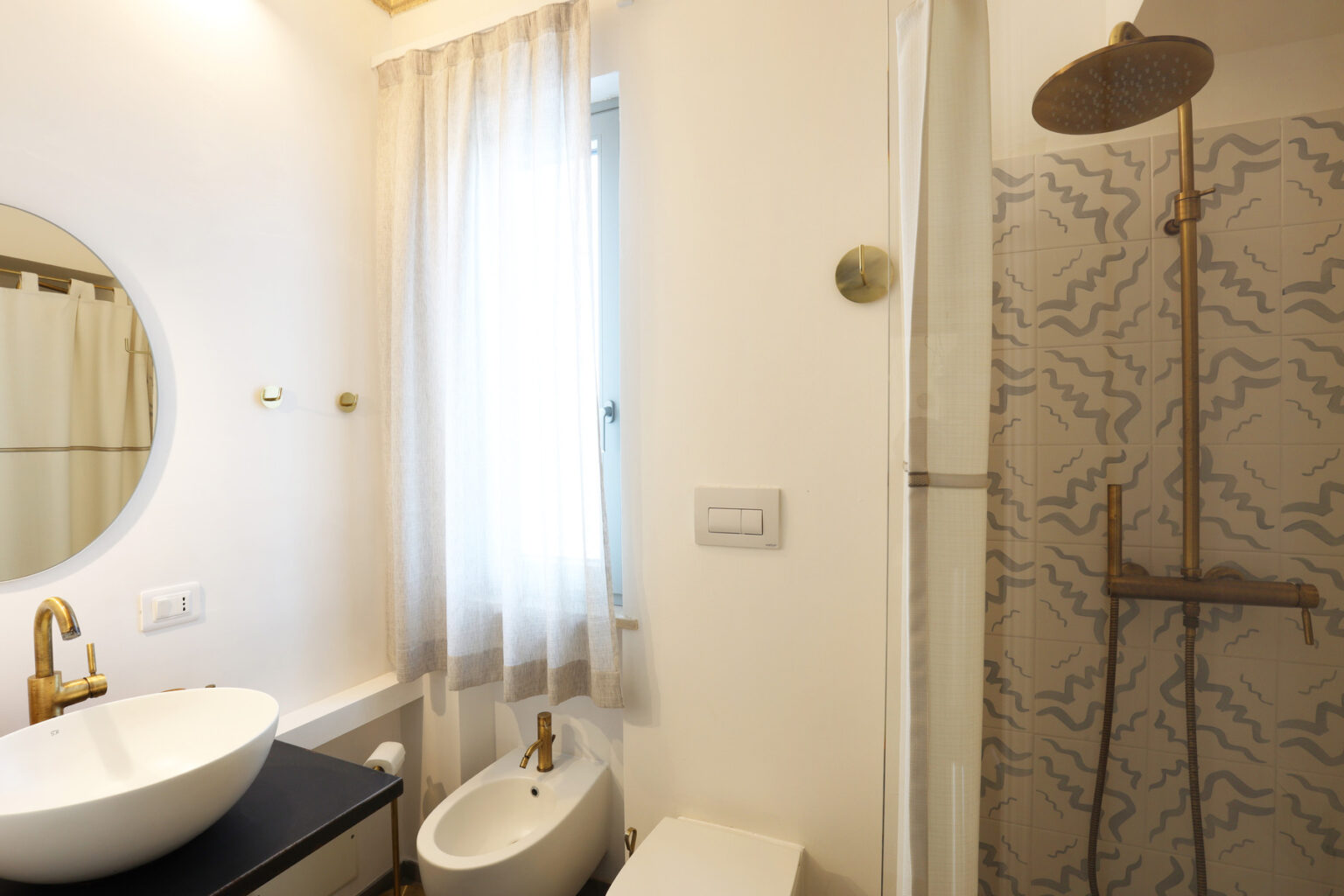 Modern bathroom featuring a circular mirror, elegant sink, and a stylish shower with a textured tile wall. Natural light filters through sheer curtains, enhancing the contemporary design. | Only on SopranoVillas
