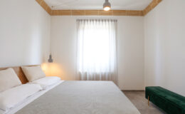 Modern bedroom interior featuring a neutral color palette, a cozy bed with light bedding, a large window with sheer curtains allowing natural light, and a stylish green bench accent piece.