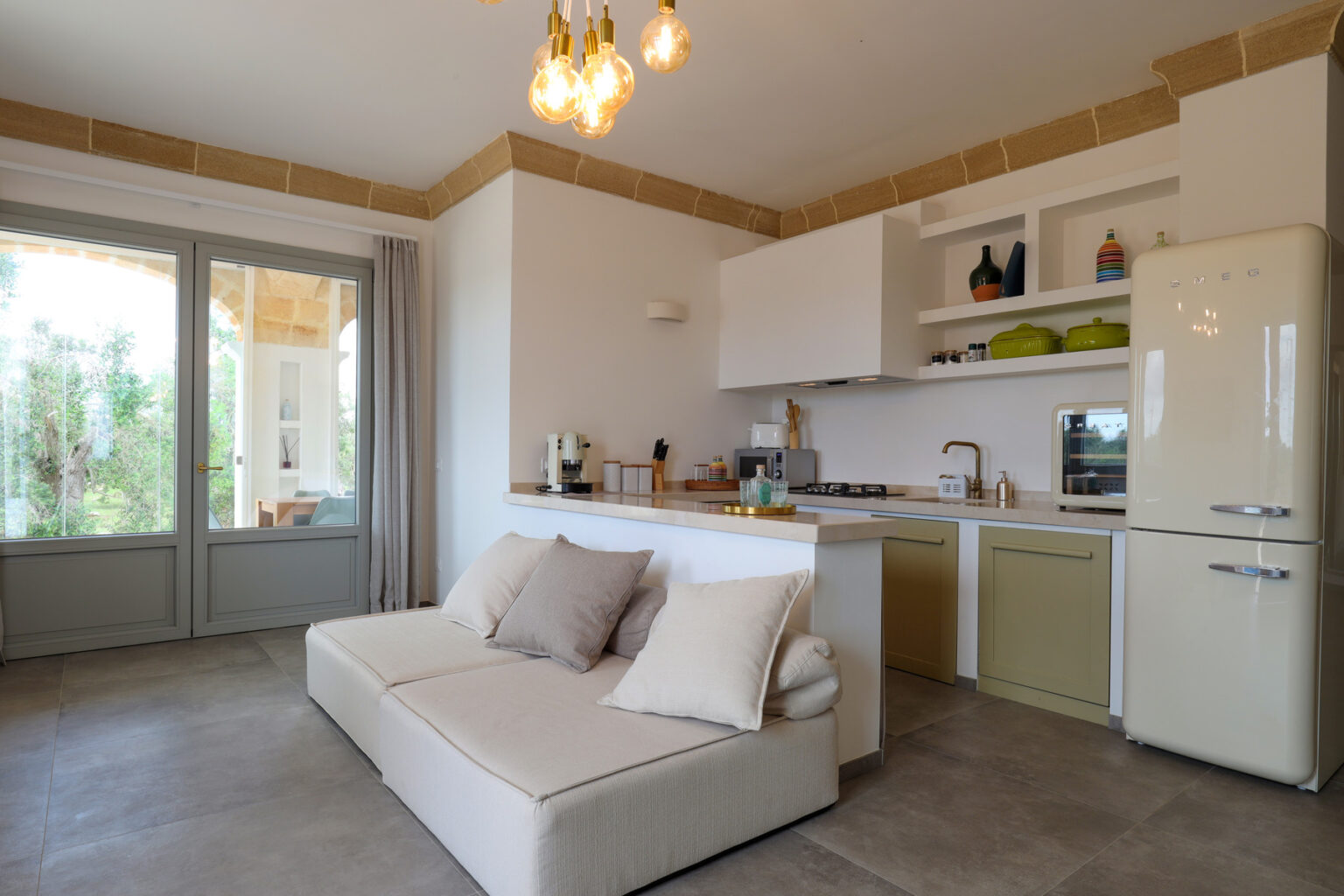 Modern kitchen and living area featuring a cozy white sofa with pillows, stylish cabinetry, and large windows that allow natural light to fill the space. The kitchen is equipped with contemporary appliances and decorative accents, creating a welcoming atmosphere. | Only on SopranoVillas