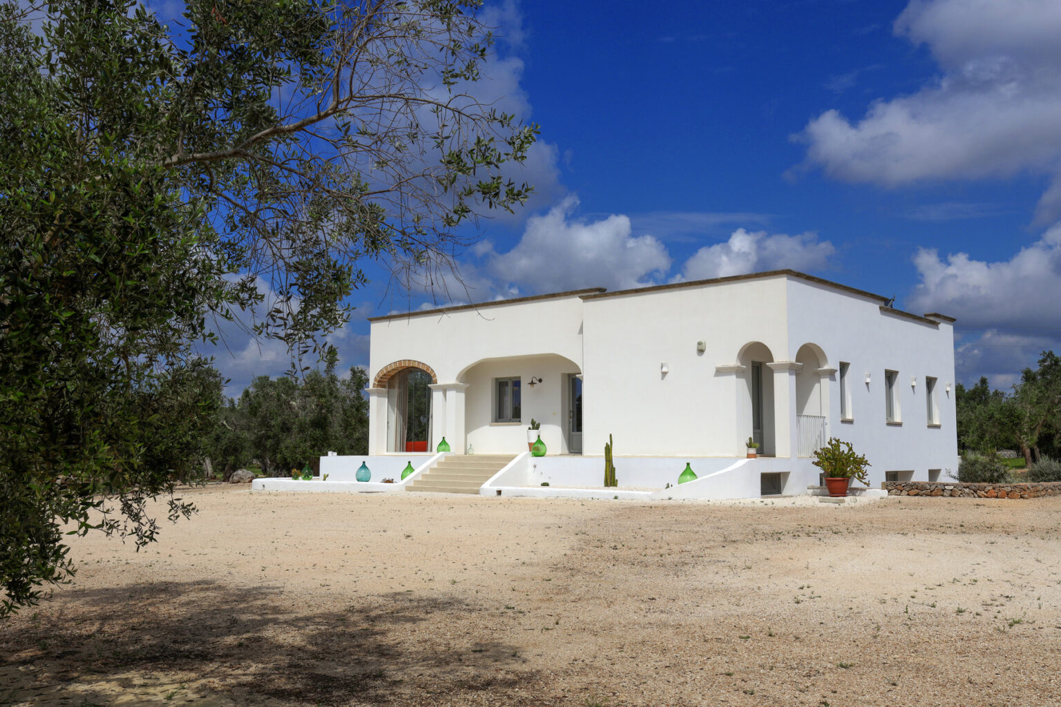 White Mediterranean-style house surrounded by olive trees under a blue sky with fluffy clouds, featuring arched windows and a spacious front porch. Ideal for showcasing rural architecture and serene landscapes. | Only on SopranoVillas