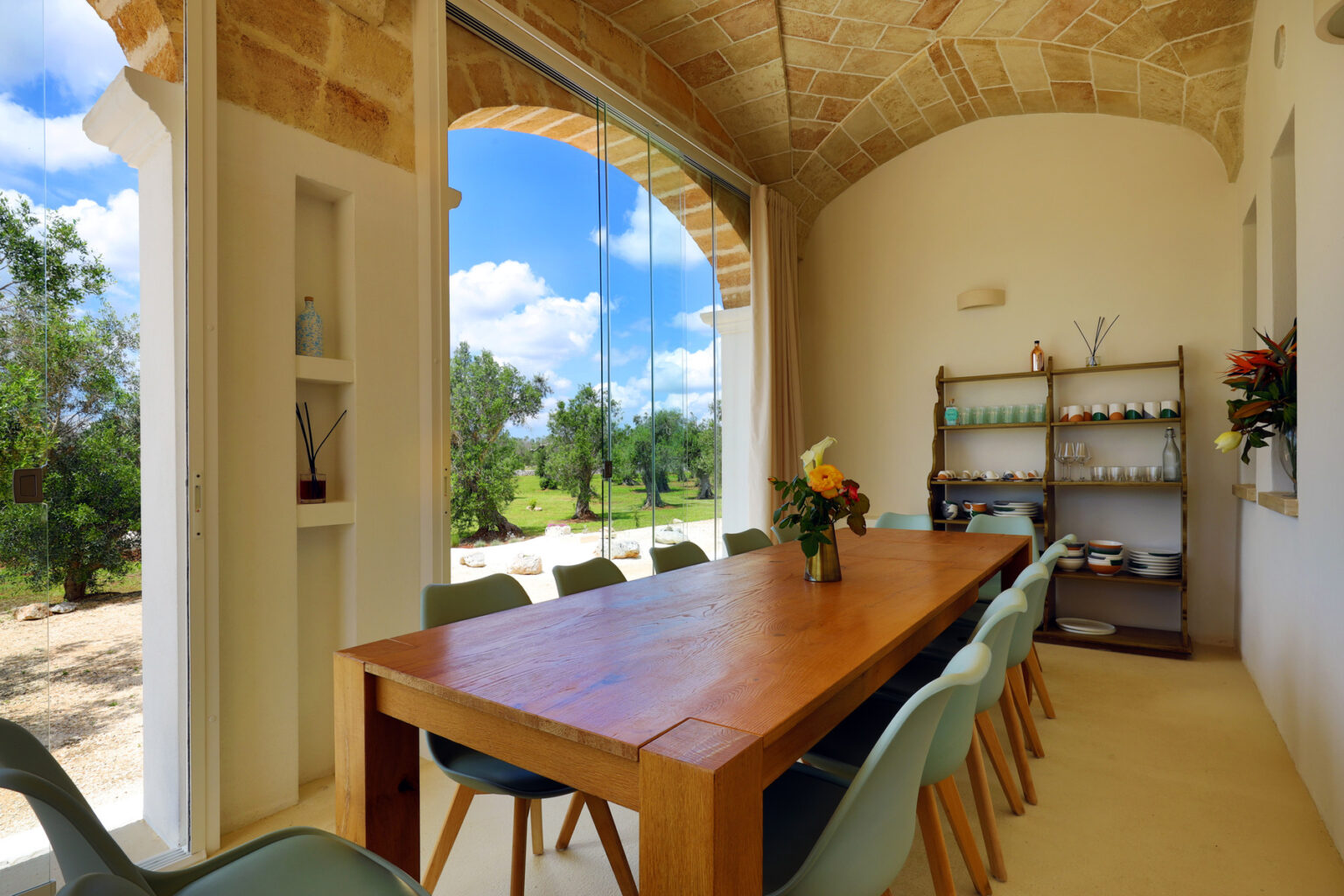 Bright and airy dining room featuring a large wooden table surrounded by light blue chairs, with large windows offering a view of the outdoors and lush greenery. The room is decorated with a small shelf displaying various items and fresh flowers, creating a welcoming atmosphere. | Only on SopranoVillas