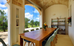 Bright and airy dining room featuring a large wooden table surrounded by light blue chairs, with large windows offering a view of the outdoors and lush greenery. The room is decorated with a small shelf displaying various items and fresh flowers, creating a welcoming atmosphere.