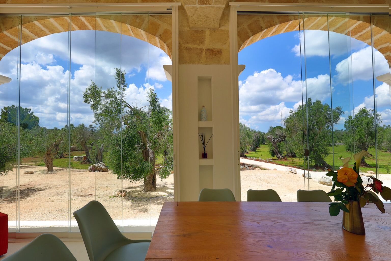 Interior view of a modern dining area with a wooden table and chairs, showcasing large glass windows that open to a scenic outdoor landscape featuring trees and blue skies with clouds. | Only on SopranoVillas