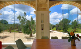 Interior view of a modern dining area with a wooden table and chairs, showcasing large glass windows that open to a scenic outdoor landscape featuring trees and blue skies with clouds.