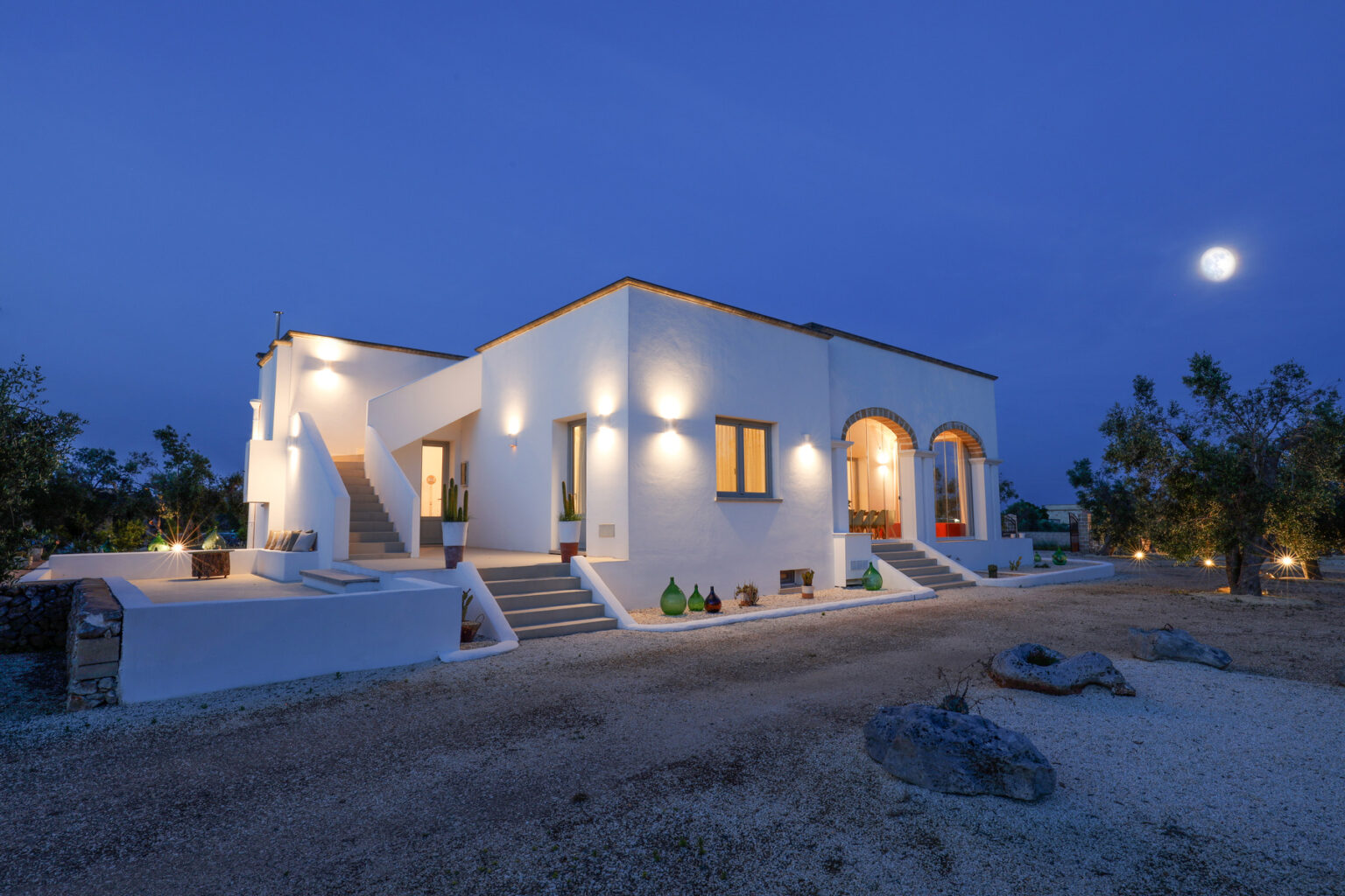 Modern white villa illuminated at night, featuring a spacious entrance and landscaped surroundings, set against a clear blue sky with a full moon. | Only on SopranoVillas