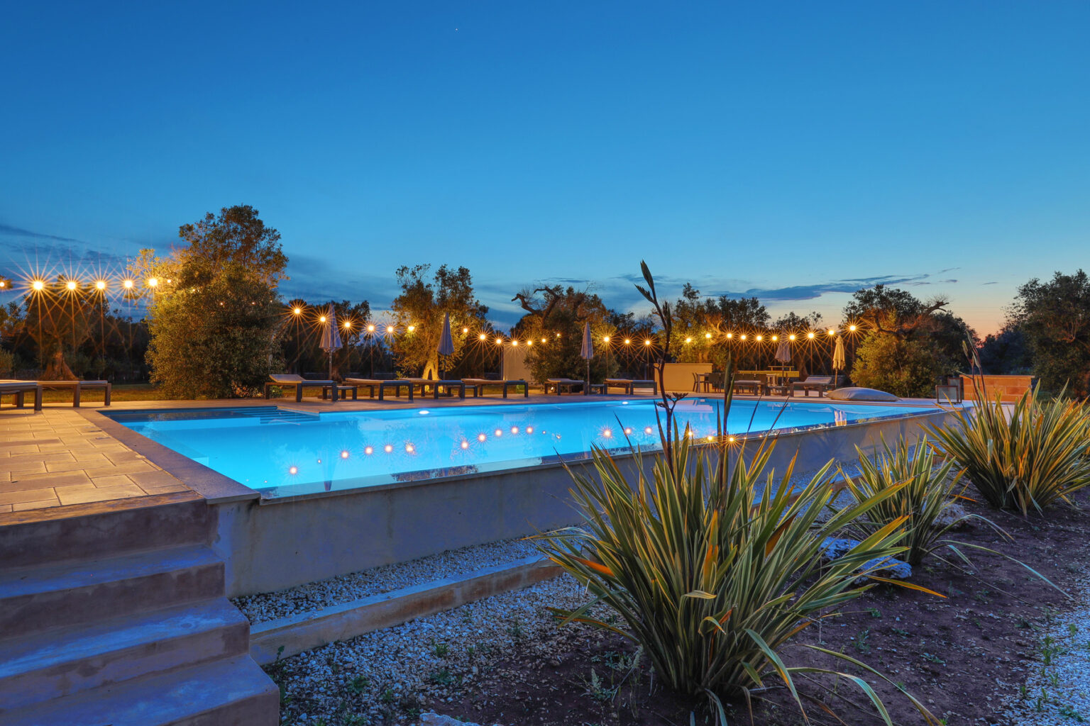 Serene outdoor swimming pool illuminated by string lights at dusk, surrounded by landscaped greenery and a peaceful atmosphere. Ideal for relaxation and evening gatherings. | Only on SopranoVillas