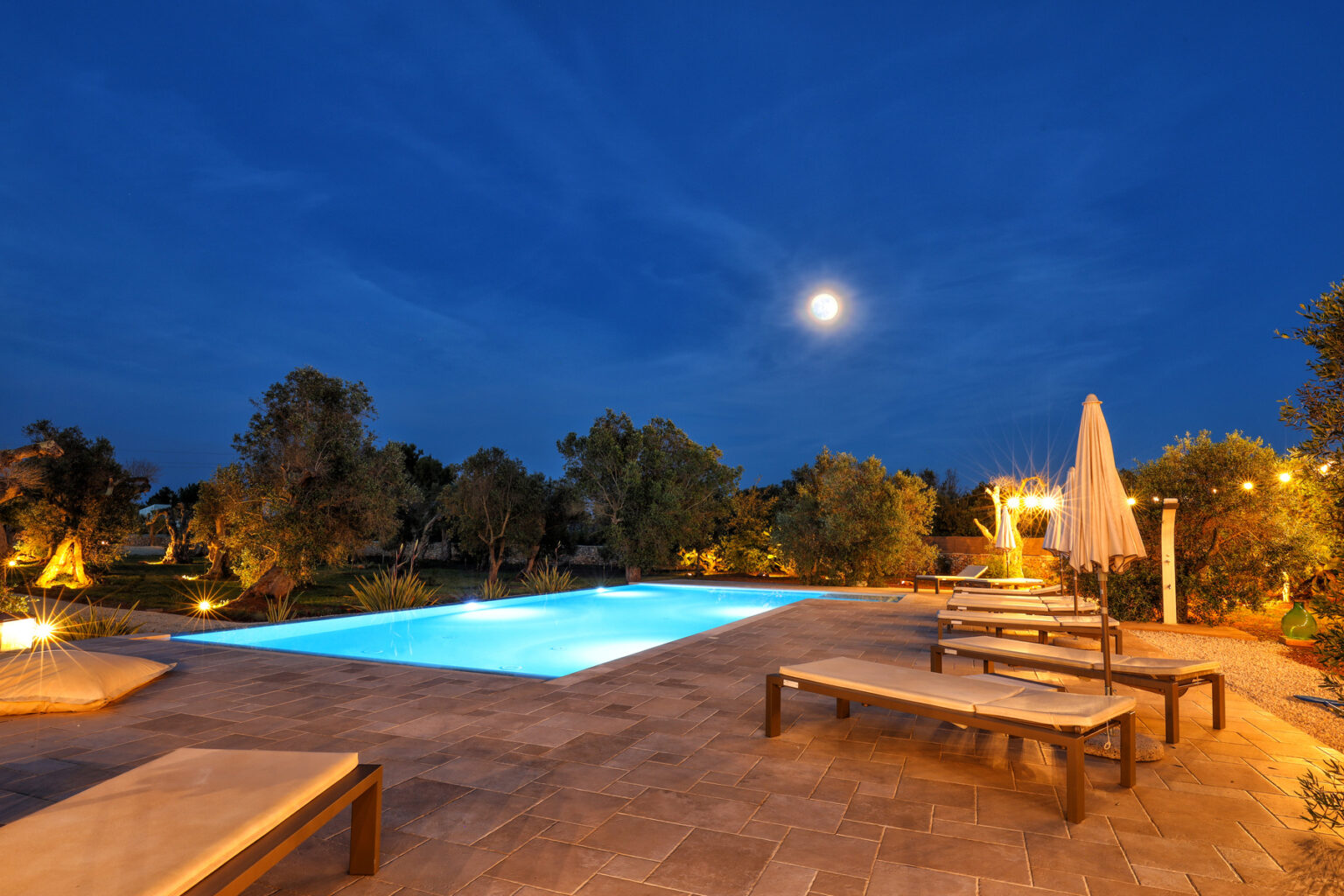 Outdoor swimming pool illuminated at night, surrounded by trees and lounge chairs, with a full moon in a clear sky. Ideal for relaxation and evening gatherings. | Only on SopranoVillas