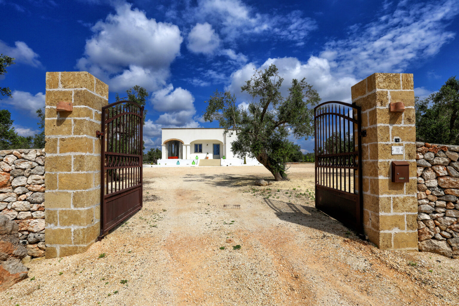 Rustic entrance gate leading to a charming white house surrounded by olive trees under a blue sky with fluffy clouds, set in a serene landscape. | Only on SopranoVillas