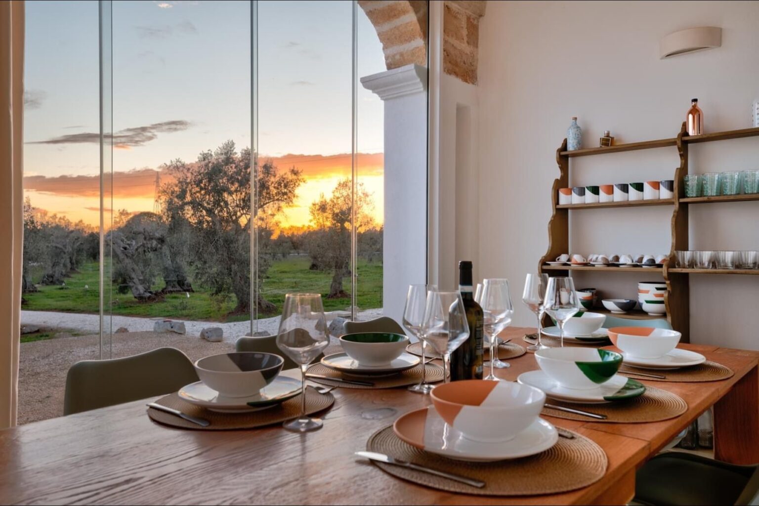 Modern dining room with a wooden table set for a meal, featuring elegant tableware and wine glasses. Large windows showcase a stunning sunset over a green landscape, creating a warm and inviting atmosphere. | Only on SopranoVillas