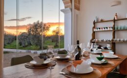Modern dining room with a wooden table set for a meal, featuring elegant tableware and wine glasses. Large windows showcase a stunning sunset over a green landscape, creating a warm and inviting atmosphere.