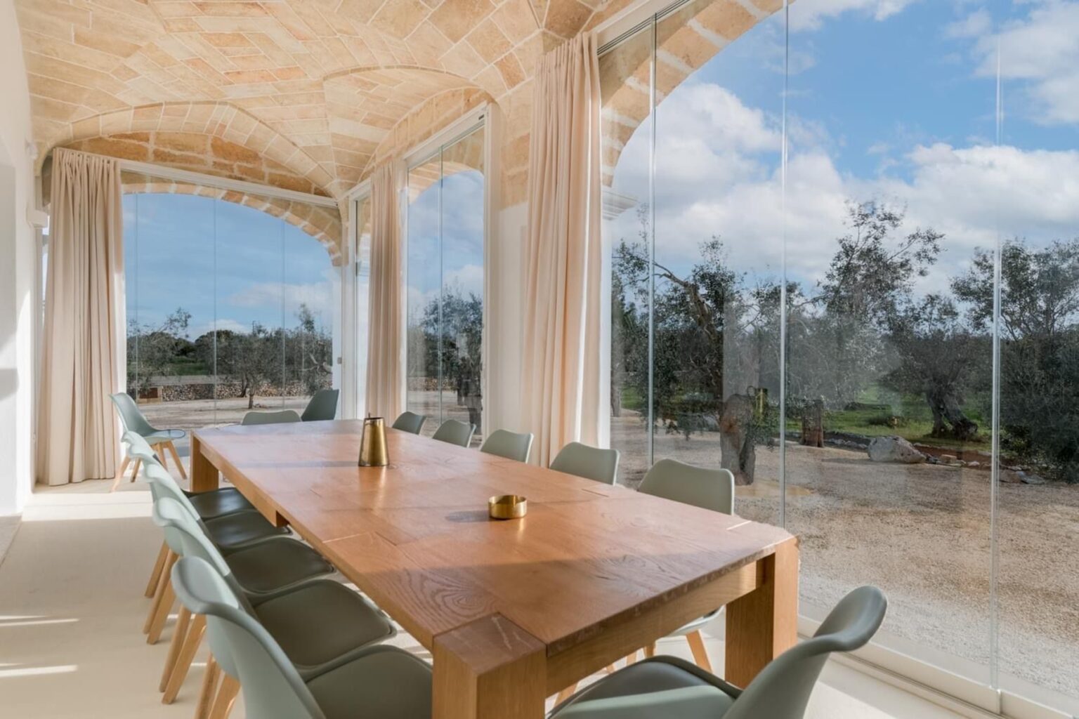 Modern dining room with a wooden table and green chairs, featuring large glass windows that offer a view of a landscaped outdoor area with trees and blue skies. Natural light floods the space, enhancing the contemporary design. | Only on SopranoVillas