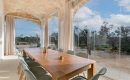 Modern dining room with a wooden table and green chairs, featuring large glass windows that offer a view of a landscaped outdoor area with trees and blue skies. Natural light floods the space, enhancing the contemporary design.