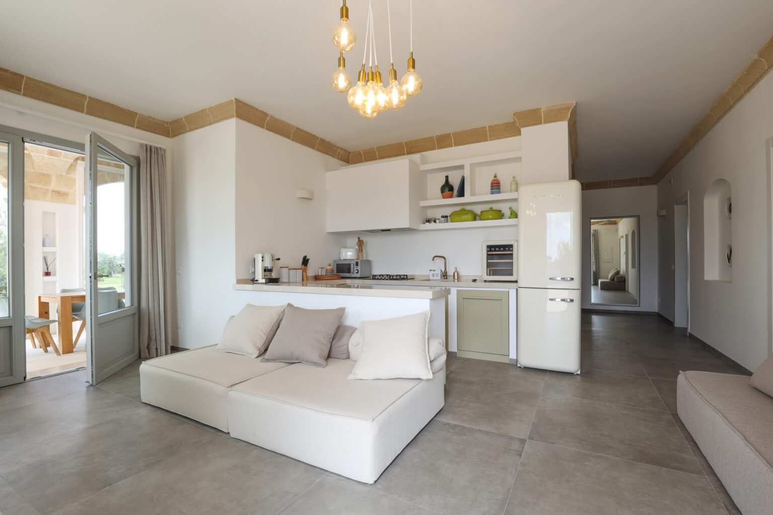 Modern open-concept living room and kitchen featuring a white sofa, minimalist decor, and large windows providing natural light. The kitchen includes sleek cabinetry and a refrigerator, with a dining area visible in the background. | Only on SopranoVillas