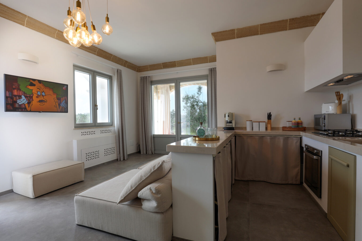Modern kitchen and living area featuring a sofa, pendant lighting, and a wall-mounted TV. The space is bright with large windows and minimalistic decor, ideal for comfortable living. | Only on SopranoVillas