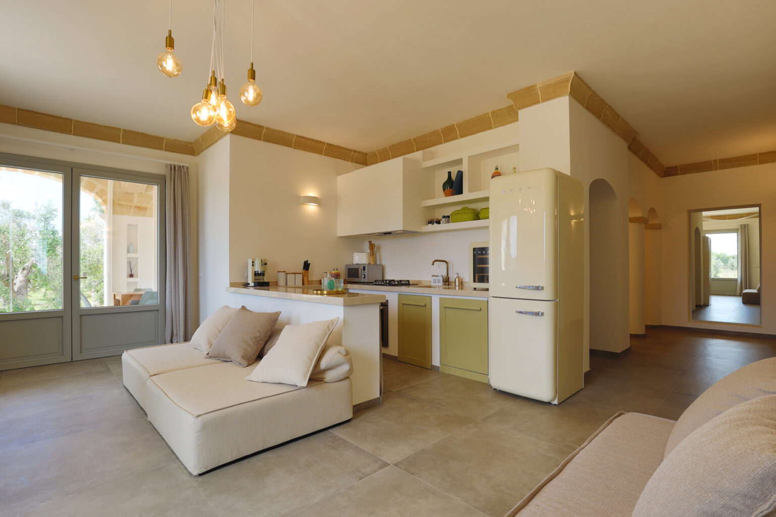 Modern living room and kitchen area featuring a light color scheme, comfortable sofa, and stylish decor. The space includes a refrigerator, kitchen counter, and large windows that provide natural light. Ideal for showcasing contemporary interior design. | Only on SopranoVillas
