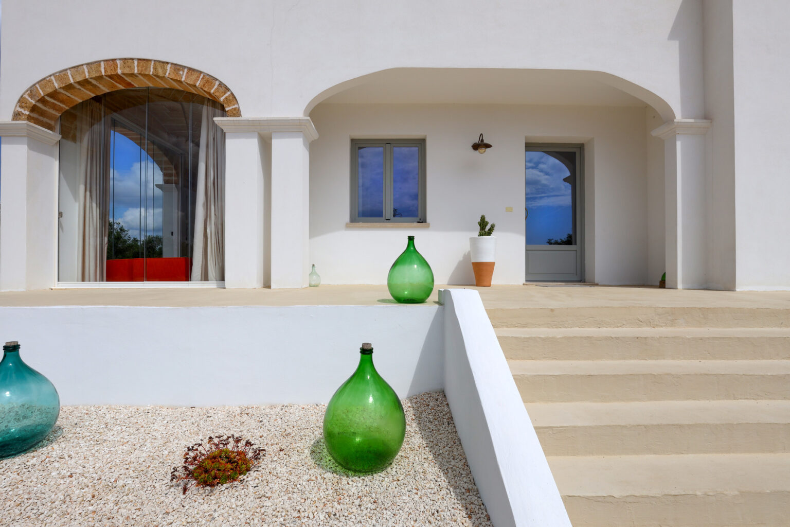 Modern entrance of a white Mediterranean-style house featuring decorative glass vases, a stone pathway, and a large window with a view of the outdoors. | Only on SopranoVillas