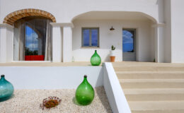 Modern entrance of a white Mediterranean-style house featuring decorative glass vases, a stone pathway, and a large window with a view of the outdoors.