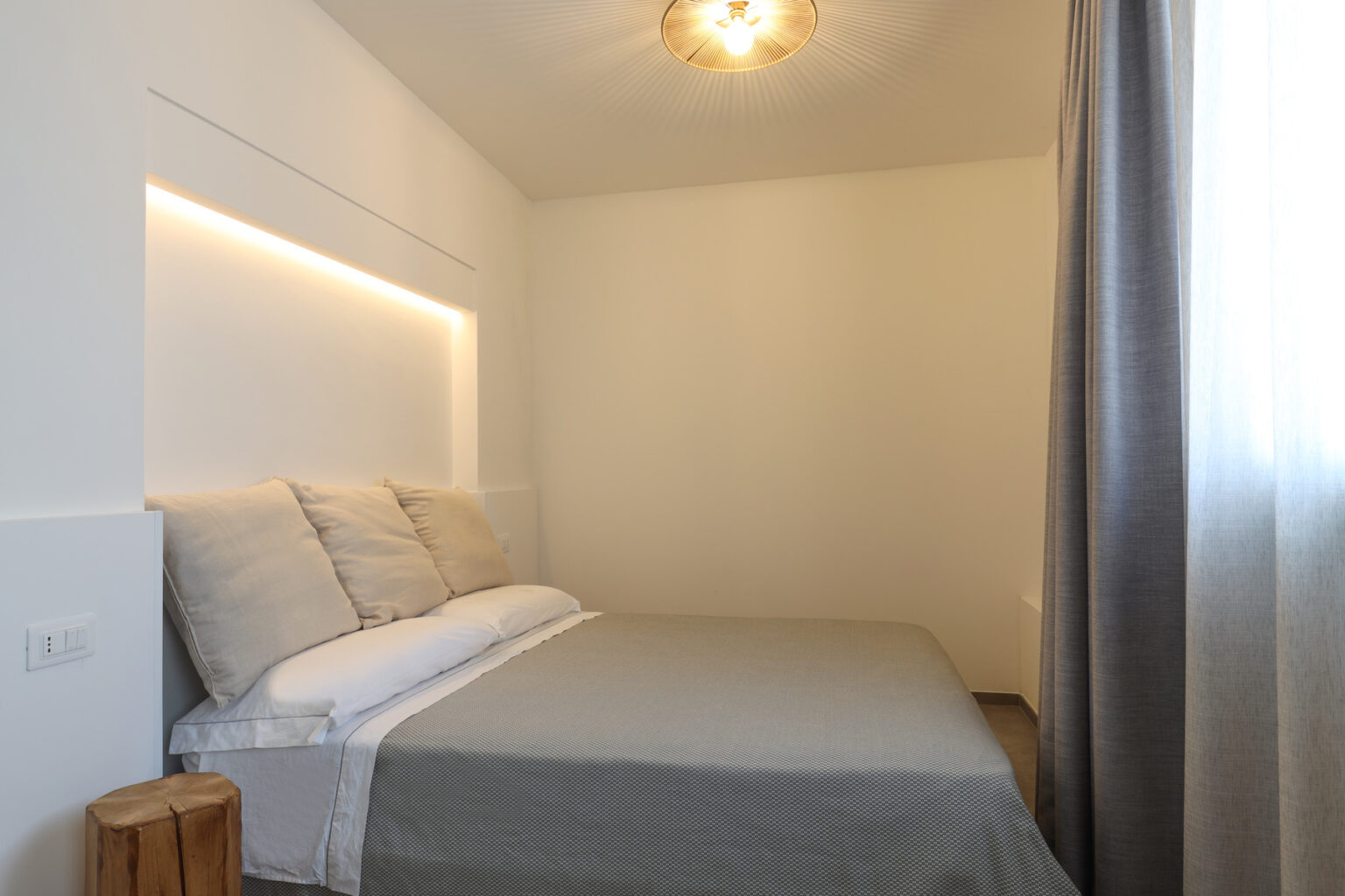 Modern bedroom interior featuring a comfortable bed with light-colored pillows and a gray blanket. The room is illuminated by soft LED lighting above the headboard, and a stylish pendant light hangs from the ceiling. Natural light filters through sheer curtains, enhancing the minimalist design and serene atmosphere. | Only on SopranoVillas