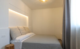 Modern bedroom interior featuring a comfortable bed with light-colored pillows and a gray blanket. The room is illuminated by soft LED lighting above the headboard, and a stylish pendant light hangs from the ceiling. Natural light filters through sheer curtains, enhancing the minimalist design and serene atmosphere.