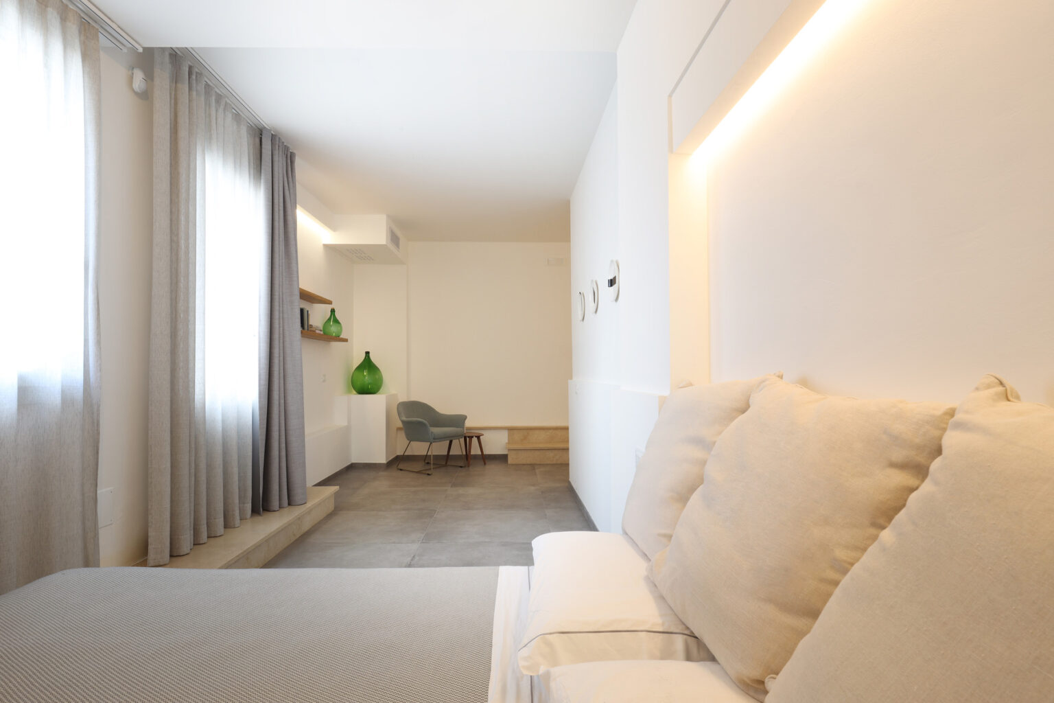Modern hotel room featuring a cozy bed with soft pillows, light gray curtains, and a minimalistic design. A green decorative vase and a stylish chair add a pop of color to the space, while natural light illuminates the room, enhancing its contemporary aesthetic. | Only on SopranoVillas