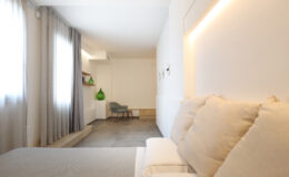 Modern hotel room featuring a cozy bed with soft pillows, light gray curtains, and a minimalistic design. A green decorative vase and a stylish chair add a pop of color to the space, while natural light illuminates the room, enhancing its contemporary aesthetic.