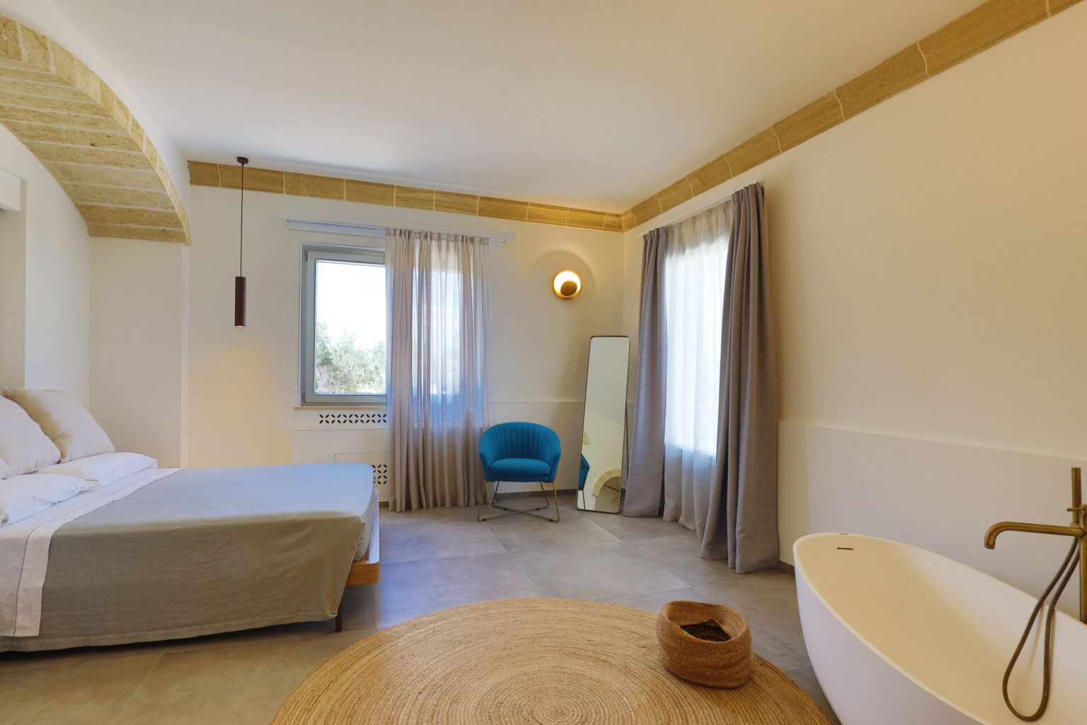 Modern bedroom interior featuring a comfortable bed, minimalist decor, a stylish blue chair, and a freestanding bathtub. Natural light flows through large windows, complemented by soft curtains and a decorative round rug on the floor. | Only on SopranoVillas