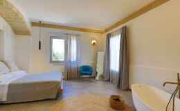 Modern bedroom interior featuring a comfortable bed, minimalist decor, a stylish blue chair, and a freestanding bathtub. Natural light flows through large windows, complemented by soft curtains and a decorative round rug on the floor.