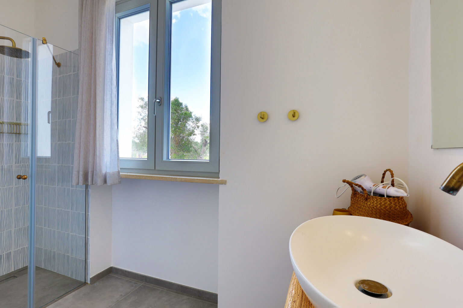 Modern bathroom interior featuring a glass shower, large window with natural light, and stylish sink with a woven basket for storage. | Only on SopranoVillas