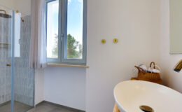 Modern bathroom interior featuring a glass shower, large window with natural light, and stylish sink with a woven basket for storage.