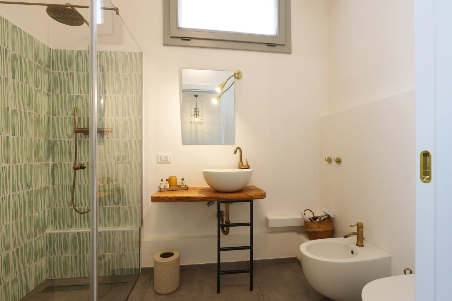 Modern bathroom featuring a glass shower with green patterned tiles, a wooden vanity with a round sink, stylish brass fixtures, and minimalist decor. Natural light enters through a window, enhancing the bright and airy atmosphere. | Only on SopranoVillas