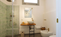 Modern bathroom featuring a glass shower with green patterned tiles, a wooden vanity with a round sink, stylish brass fixtures, and minimalist decor. Natural light enters through a window, enhancing the bright and airy atmosphere.