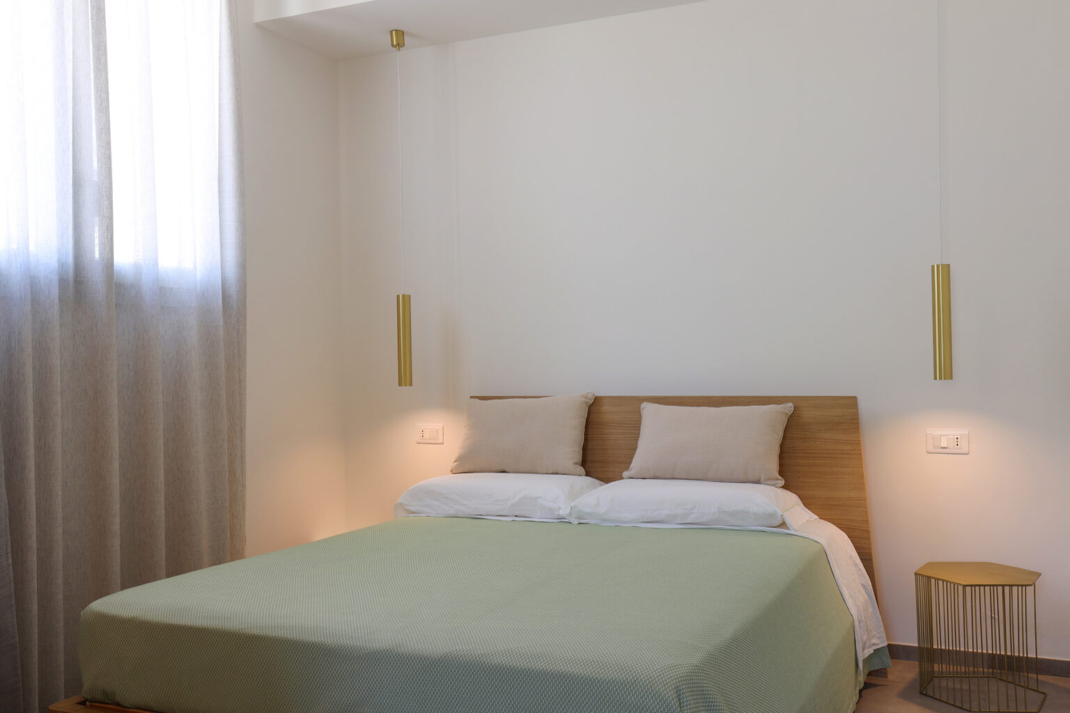Modern bedroom interior featuring a queen-sized bed with a light green blanket, wooden headboard, and bedside lamps, complemented by sheer curtains and minimalist decor. | Only on SopranoVillas