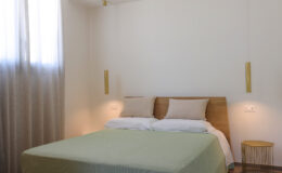 Modern bedroom interior featuring a queen-sized bed with a light green blanket, wooden headboard, and bedside lamps, complemented by sheer curtains and minimalist decor.