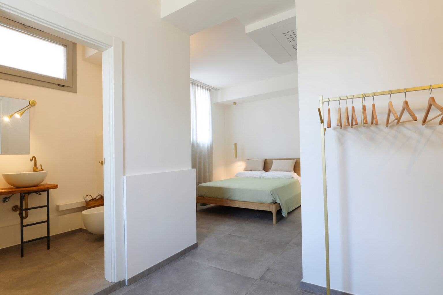 Modern interior of a stylish bedroom with a green bedspread, featuring a connected bathroom and a wooden coat rack. Natural light fills the space, highlighting the minimalist design and neutral color palette. | Only on SopranoVillas