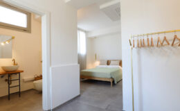 Modern interior of a stylish bedroom with a green bedspread, featuring a connected bathroom and a wooden coat rack. Natural light fills the space, highlighting the minimalist design and neutral color palette.