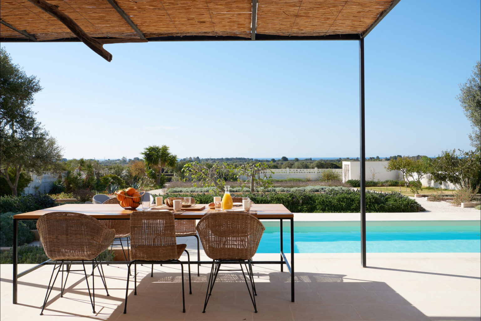 Outdoor dining area with a modern table and woven chairs, overlooking a scenic view of a swimming pool and lush landscape under a clear blue sky. Ideal for relaxation and entertaining. | Only on SopranoVillas