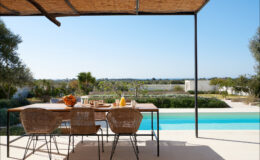 Outdoor dining area with a modern table and woven chairs, overlooking a scenic view of a swimming pool and lush landscape under a clear blue sky. Ideal for relaxation and entertaining.