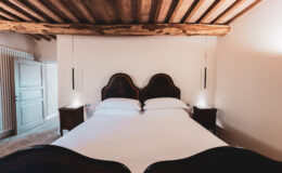Cozy bedroom featuring a double bed with elegant wooden headboards, soft white linens, and stylish bedside tables. The room showcases rustic wooden beams and a warm color palette, creating a welcoming atmosphere. A door leads to an adjoining space, enhancing the room's charm. Ideal for a relaxing getaway.