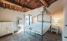 Elegant bedroom featuring a four-poster bed with a canopy, wooden ceiling beams, and stylish furnishings including a vintage dresser and nightstand. Natural light floods the space, creating a warm and inviting atmosphere.