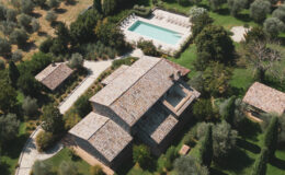 Aerial view of a luxurious villa surrounded by olive trees, featuring a large swimming pool and elegant outdoor seating areas. The rustic stone architecture and lush landscape create a serene and picturesque setting.