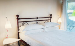Cozy bedroom featuring a vintage metal bed frame with white linens and multiple plush pillows, complemented by modern white bedside lamps and a stylish side table, with natural light streaming in from a nearby window.