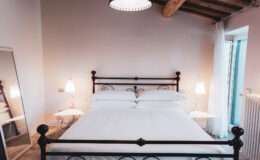 Cozy bedroom featuring a wrought iron bed with white linens, bedside lamps, and a large window allowing natural light, complemented by wooden ceiling beams and a stylish mirror.