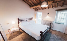 Cozy bedroom featuring a double bed with white linens, wooden ceiling beams, and natural light from a window. The room includes modern lighting and a minimalist design, creating a warm and inviting atmosphere.