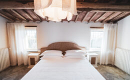 Cozy bedroom interior featuring a queen-sized bed with white linens, wooden ceiling beams, and natural light from two windows. The room includes bedside tables and soft, sheer curtains, creating a warm and inviting atmosphere.