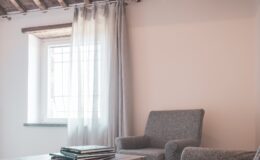 Cozy interior living space featuring two gray armchairs, a glass coffee table stacked with magazines, and sheer curtains allowing natural light to filter in through a window with wooden beams overhead.